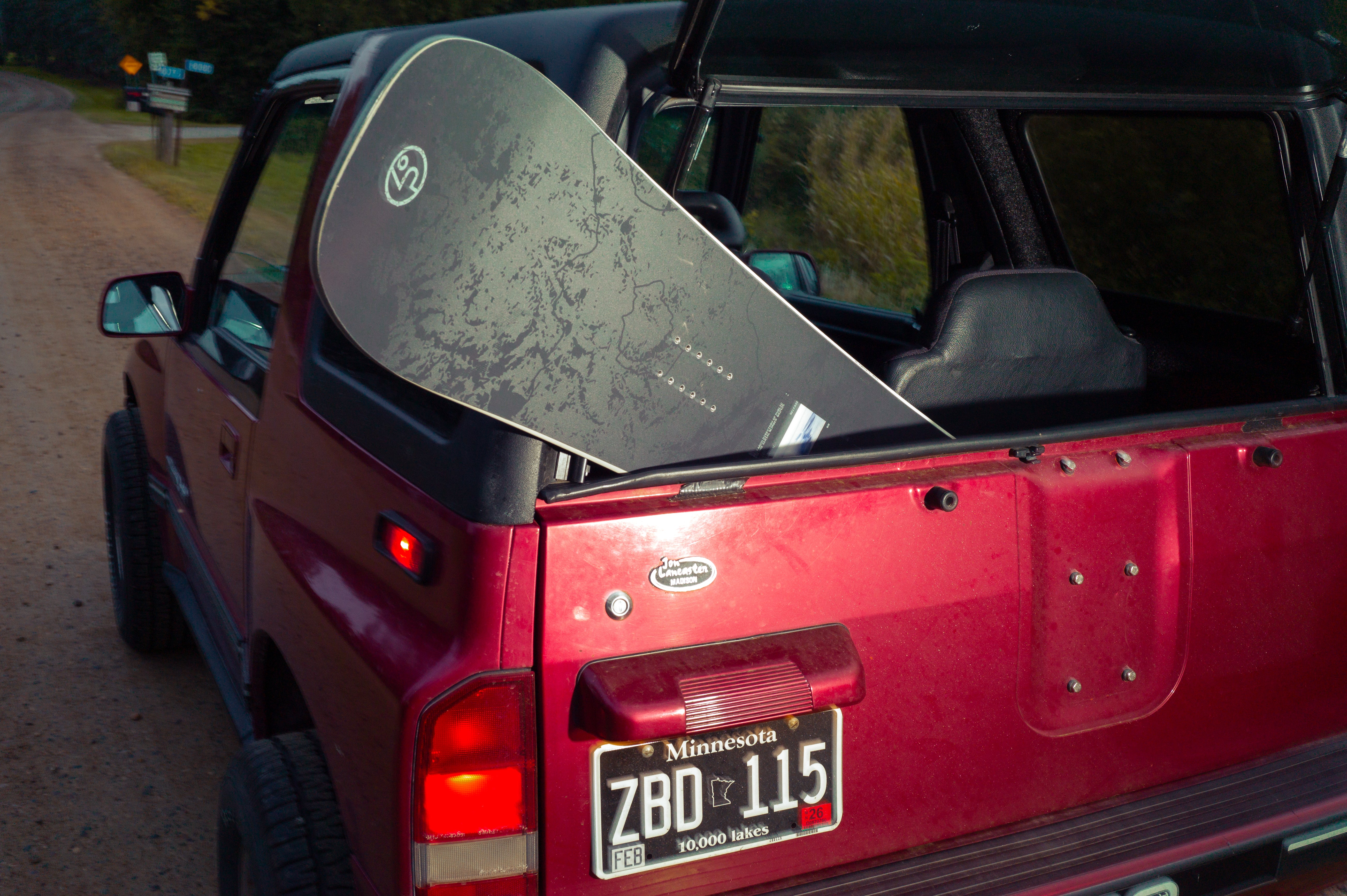 Red vehicle with a snowboard on the back, parked on a dirt road.